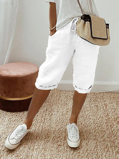 Femme portant un short blanc en lin, t-shirt gris, baskets blanches et sac à main en paille, posant sur un tapis beige. Mode décontractée estivale.