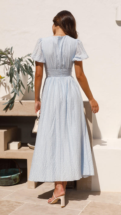 Femme en robe longue bleue à rayures, manches courtes, debout sur une terrasse ensoleillée, portant des sandales à talons, décor minimaliste.