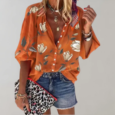Chemisier orange à fleurs blanches, manches larges, porté avec short en jean. Accessoires: collier, bracelets, pochette léopard. Mode estivale chic.