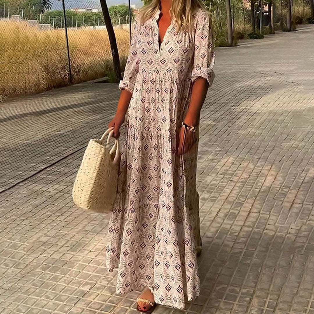 Femme en robe longue bohème imprimée, tenant un sac en osier, marchant sur un chemin pavé. Mode estivale, style décontracté chic.