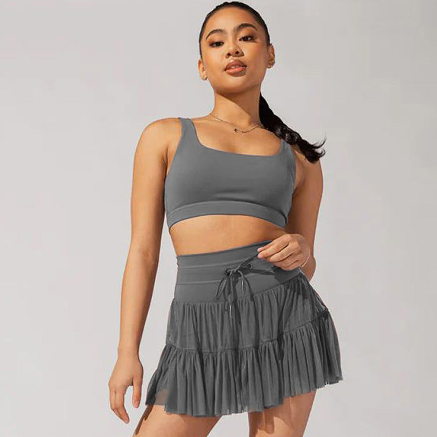 Jeune femme portant une tenue de sport grise, composée d'un haut court et d'une jupe plissée, posant sur fond neutre. Mode fitness tendance.