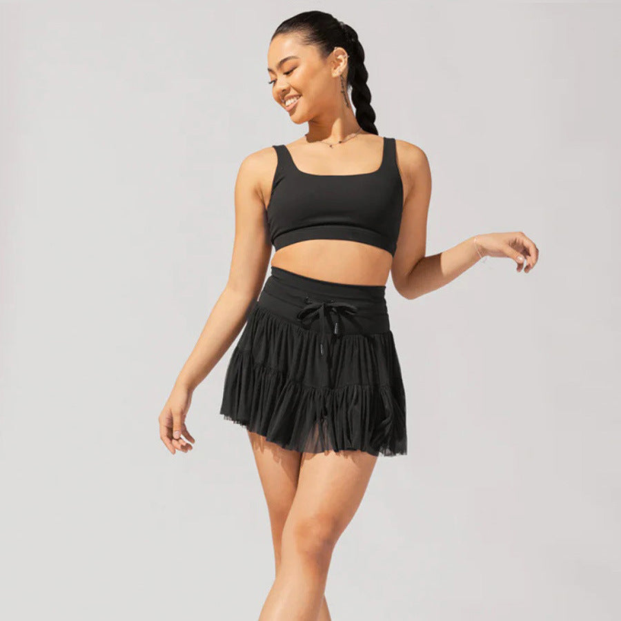 Femme souriante en tenue de sport noire, portant un haut court et une jupe plissée, posant sur fond neutre. Mode active, style décontracté.