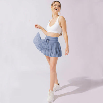Jeune femme souriante en tenue de sport, portant un haut blanc et une jupe plissée bleue, posant sur fond clair. Mode fitness féminine tendance.