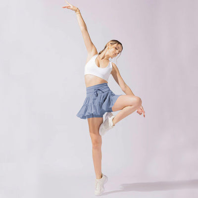 Jeune femme en tenue de sport, brassière blanche et jupe bleue, effectuant une pose de danse dynamique sur fond neutre. Fitness, danse, mode active.