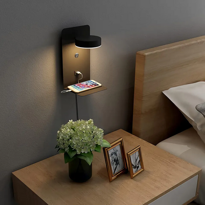 Lampe murale moderne avec étagère intégrée, chargeur USB, smartphone posé, table de chevet en bois, vase de fleurs, cadres photo, chambre élégante.