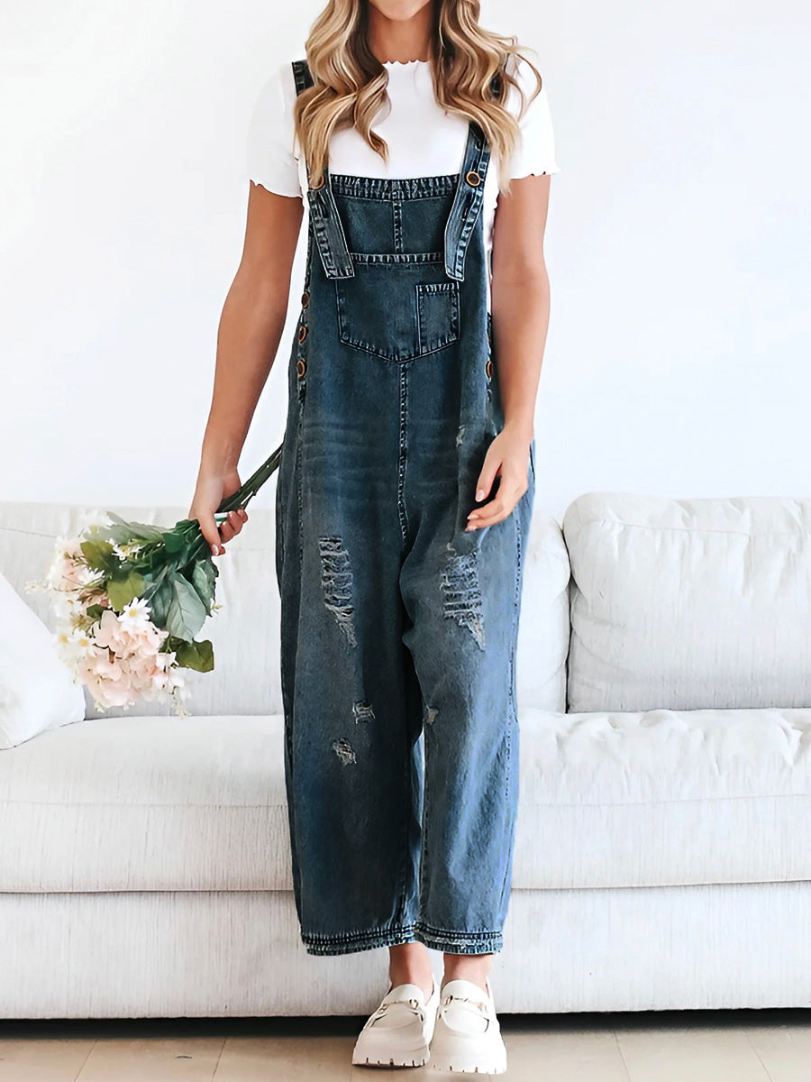 Femme en salopette en jean décontractée, tenant un bouquet de fleurs, debout devant un canapé blanc. Mode tendance, style décontracté.