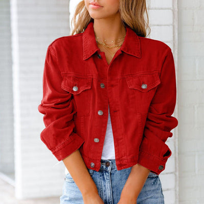 Femme portant une veste en jean rouge, manches retroussées, sur un t-shirt blanc, avec un collier doré, devant un mur blanc. Mode décontractée tendance.