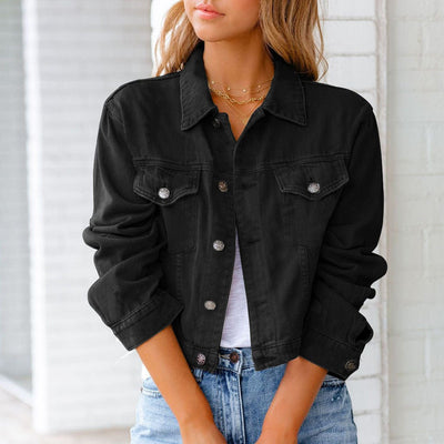 Femme portant une veste en jean noire, manches retroussées, sur un t-shirt blanc, avec un collier doré, devant un mur blanc. Mode décontractée chic.
