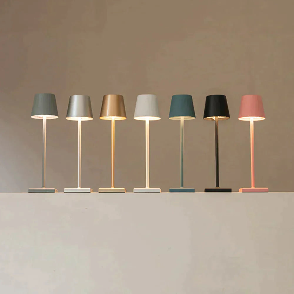 Sept lampes de table modernes alignées, chacune d'une couleur différente : gris, argent, or, beige, blanc, noir et rose, sur fond neutre.