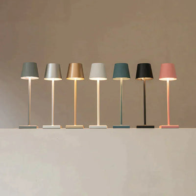 Sept lampes de table modernes alignées, chacune d'une couleur différente : gris, argent, or, beige, blanc, noir et rose, sur fond neutre.