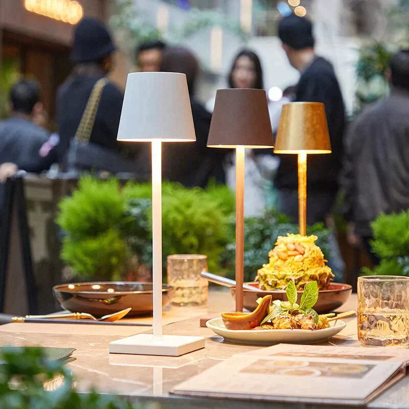 Trois lampes de table modernes éclairent un repas élégant sur une table de restaurant, avec des plats raffinés et des boissons, ambiance conviviale en arrière-plan.