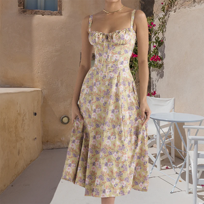 Robe d'été fleurie jaune et violette, style vintage, portée par une femme sur une terrasse ensoleillée avec chaises blanches et mur en pierre.