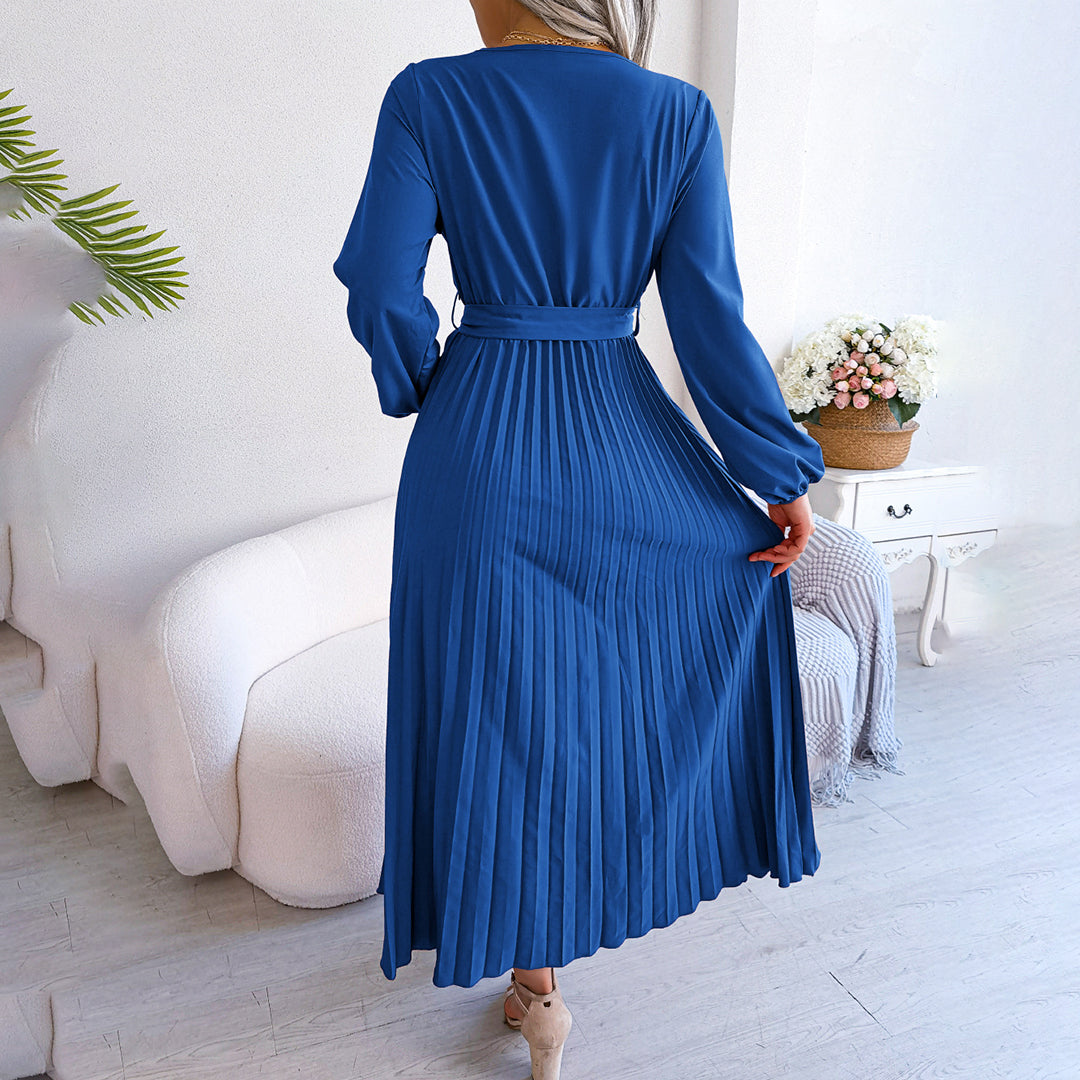 Robe longue plissée bleue élégante, manches longues, ceinture à la taille, posée dans un salon moderne avec canapé blanc et plante verte.