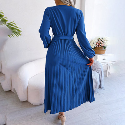 Robe longue plissée bleue élégante, manches longues, ceinture à la taille, posée dans un salon moderne avec canapé blanc et plante verte.