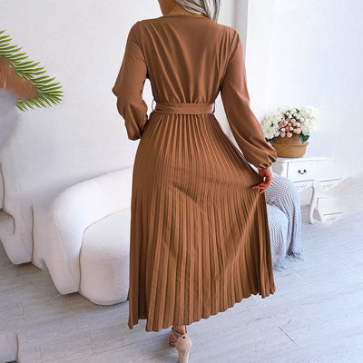 Robe plissée marron élégante portée par une femme, intérieur moderne avec canapé blanc et plante verte, ambiance chic et décontractée.