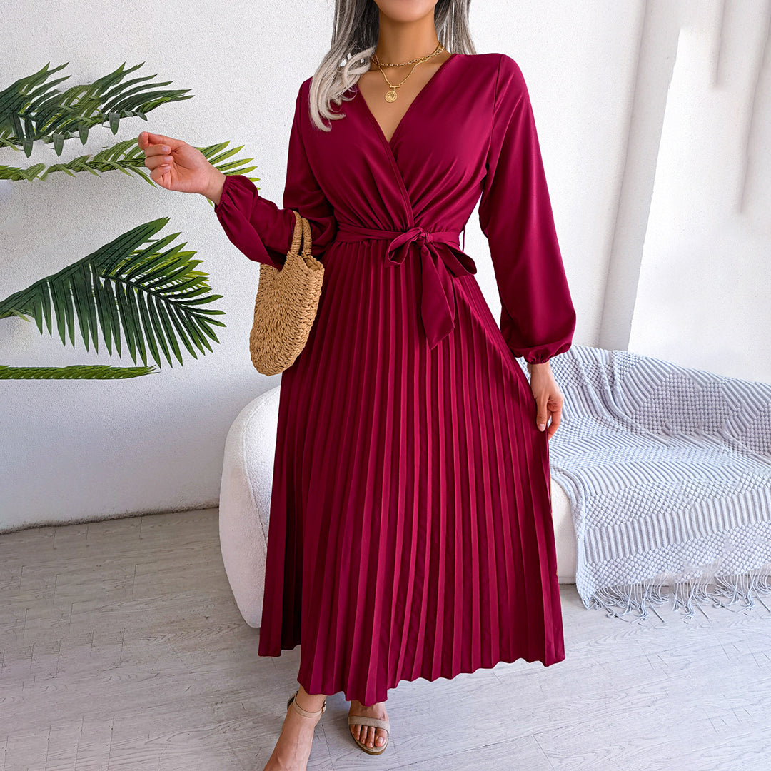 Robe longue plissée bordeaux avec ceinture nouée, manches longues, portée par une femme. Accessoire: sac en osier. Décor: plante verte, canapé.
