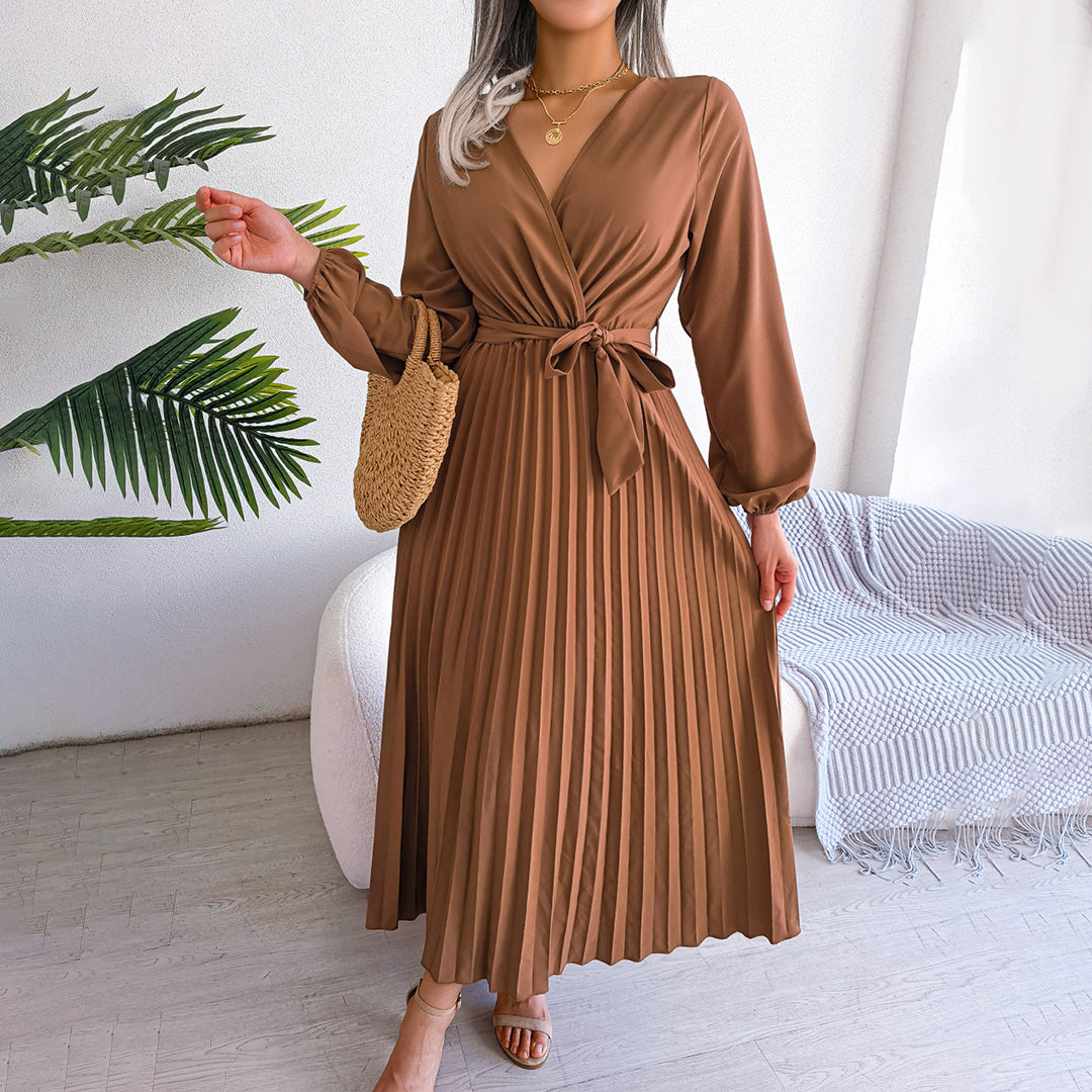 Robe longue plissée marron avec ceinture, manches longues, portée par une femme. Sac en paille à la main, décor minimaliste avec plante verte.