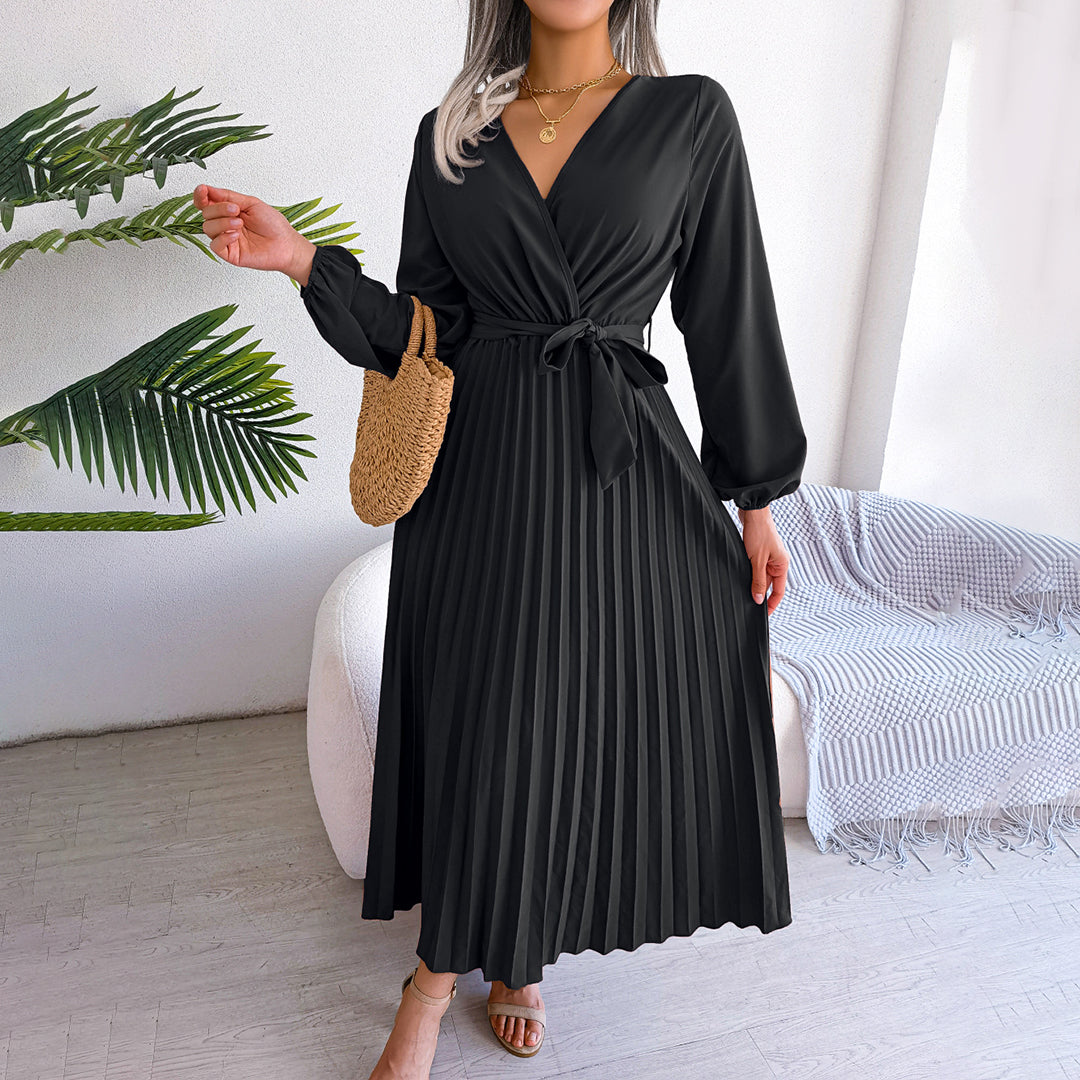 Robe noire plissée élégante avec manches longues, ceinture nouée à la taille, portée par une femme tenant un sac en osier, décor moderne en arrière-plan.