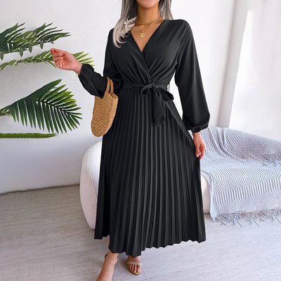 Robe noire plissée élégante avec manches longues, ceinture nouée à la taille, portée par une femme tenant un sac en osier, décor moderne en arrière-plan.