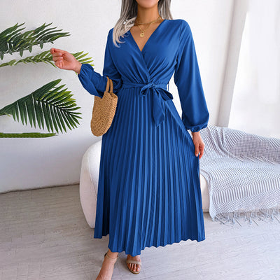 Robe bleue élégante à manches longues et ceinture nouée, posée sur un fond intérieur moderne avec plante verte et canapé. Mode féminine chic.
