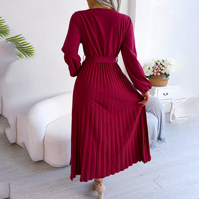 Robe longue plissée bordeaux élégante, manches longues, ceinture à la taille, posée dans un salon moderne avec canapé blanc et plante verte.