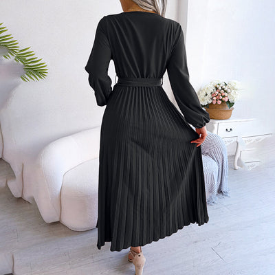 Robe longue noire plissée élégante, manches longues, ceinture à la taille, posée dans un salon moderne avec canapé blanc et plante verte.