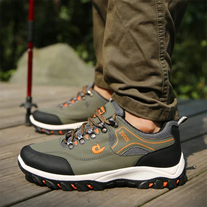 Chaussures de randonnée vertes pour homme sur plancher en bois, avec semelles antidérapantes et lacets orange, idéales pour activités de plein air.
