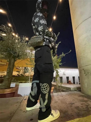 Jeune homme en tenue urbaine, portant un sweat à capuche imprimé et un pantalon cargo noir, posant sous des lumières extérieures la nuit.