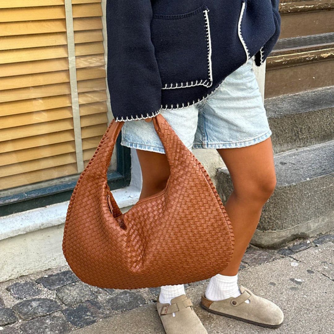 Sac à main en cuir tressé marron porté par une personne en short en jean et veste noire, devant une fenêtre avec stores en bois.
