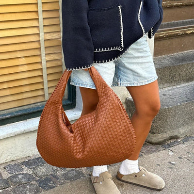 Sac à main en cuir tressé marron porté par une personne en short en jean et veste noire, devant une fenêtre avec stores en bois.