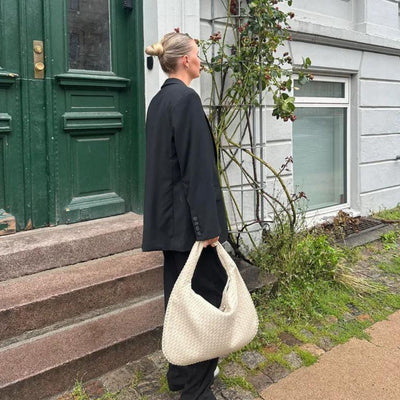 Femme en tailleur noir tenant un sac à main tissé blanc devant une maison, près d'une porte verte. Mode élégante, style urbain chic.