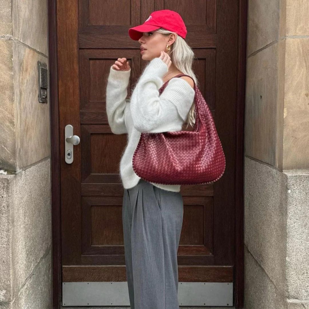 Femme élégante portant un pull blanc, pantalon gris, casquette rouge et sac à main en cuir tressé rouge, posant devant une porte en bois.