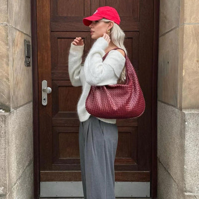 Femme élégante portant un pull blanc, pantalon gris, casquette rouge et sac à main en cuir tressé rouge, posant devant une porte en bois.