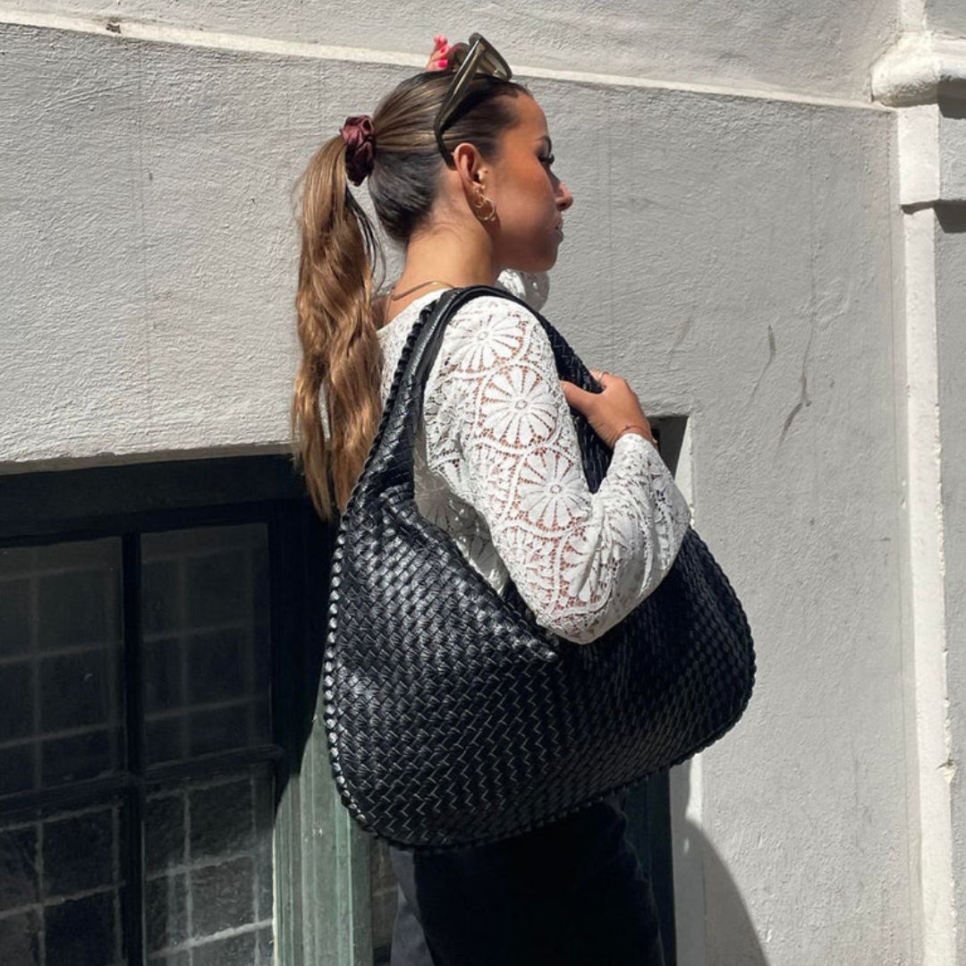 Femme portant un grand sac à main noir tressé, vêtue d'un chemisier blanc en dentelle, posant devant un mur blanc. Mode féminine, accessoires tendance.