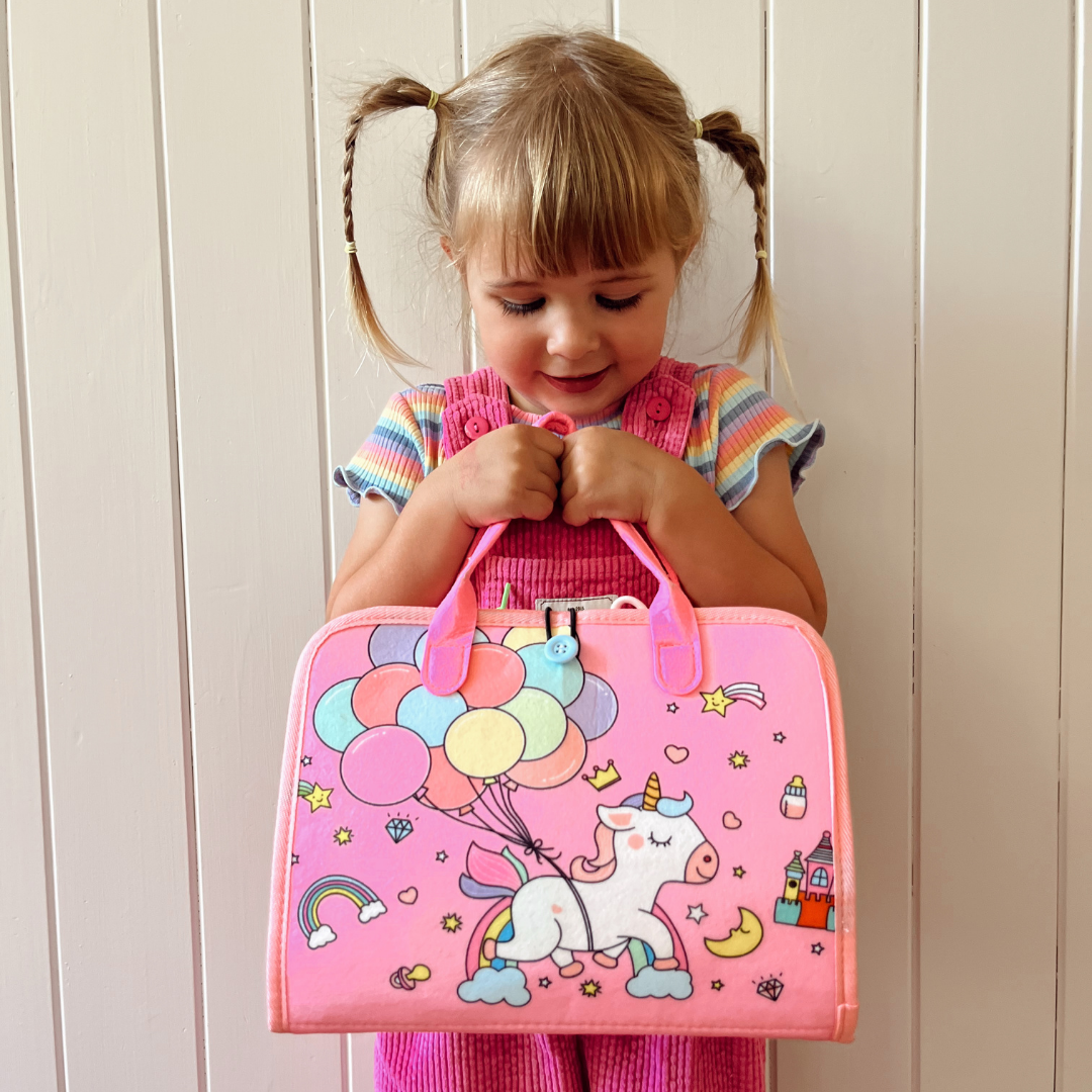 Petite fille souriante tenant une valise rose avec motif licorne et ballons colorés, devant un mur blanc. Mode enfant, accessoires licorne.