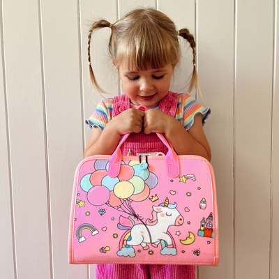 Petite fille souriante tenant une valise rose avec motif licorne et ballons colorés, devant un mur blanc. Mode enfant, accessoires licorne.