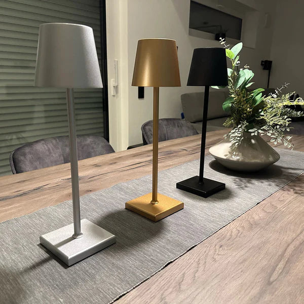 Trois lampes de table modernes en métal argenté, doré et noir sur une table en bois, avec un vase de fleurs vertes en arrière-plan. Éclairage intérieur design.