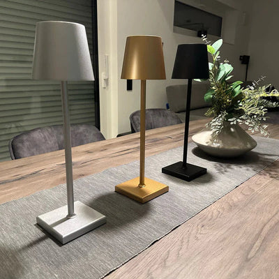 Trois lampes de table modernes en métal argenté, doré et noir sur une table en bois, avec un vase de fleurs vertes en arrière-plan. Éclairage intérieur design.