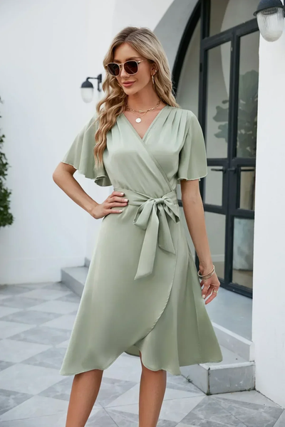 Femme portant une robe portefeuille verte élégante, manches courtes, avec lunettes de soleil, posant devant une maison moderne. Mode féminine chic.
