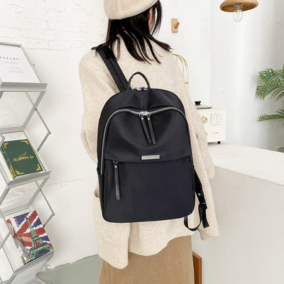 Femme de dos portant un sac à dos noir élégant, intérieur moderne, étagère avec livres, plante verte, ambiance minimaliste.