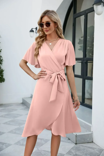 Robe portefeuille rose élégante portée par une femme, avec manches courtes et ceinture nouée, devant une maison moderne. Mode féminine chic.