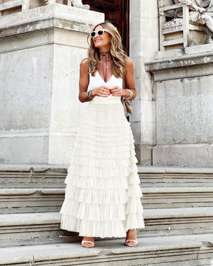 Femme élégante en jupe longue plissée blanche et débardeur blanc, posant sur des marches en pierre, lunettes de soleil rondes, style chic urbain.