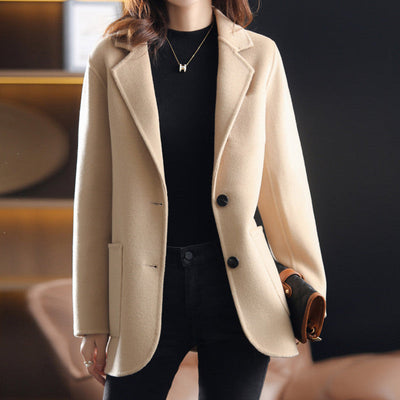 Femme portant un manteau beige élégant, col tailleur, boutons noirs, tenue chic automne-hiver, mode féminine moderne, intérieur sophistiqué.