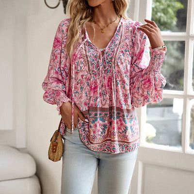 Femme portant un chemisier bohème rose à motifs floraux, manches longues, avec un sac en osier, posant près d'une fenêtre lumineuse.