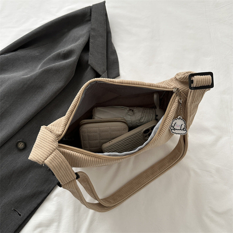 Sac en velours côtelé beige ouvert, contenant un portefeuille, un téléphone et des écouteurs, posé sur une chemise grise. Accessoire mode tendance.