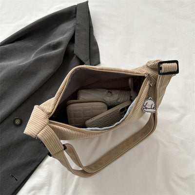 Sac en velours côtelé beige ouvert, contenant un portefeuille, un téléphone et des écouteurs, posé sur une chemise grise. Accessoire mode tendance.