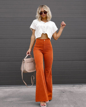 Femme en pull blanc court et pantalon orange évasé, portant un sac beige, posant devant un mur gris. Mode automne, style chic et décontracté.