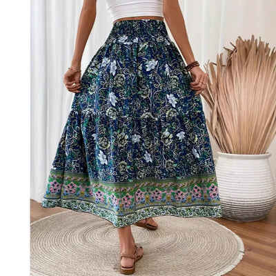 Femme portant une jupe longue bohème à motifs floraux bleus et verts, debout sur un tapis rond, décor intérieur avec vase blanc et plantes séchées.