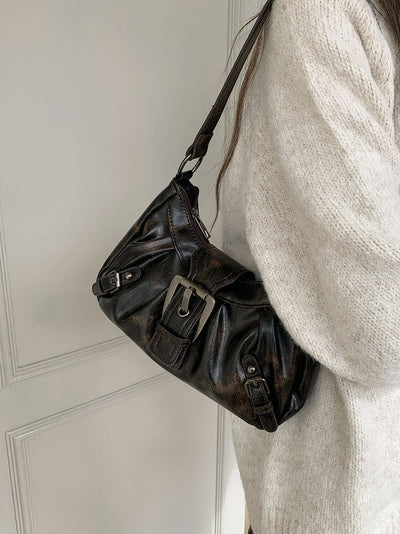 Sac à main en cuir noir avec boucle métallique, porté à l'épaule par une femme en pull beige. Mode élégante, accessoire tendance.