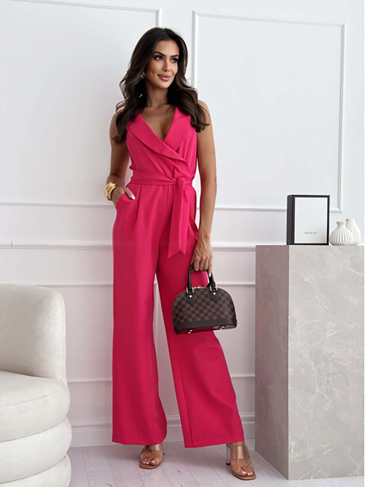 Femme portant une combinaison rose élégante, posant dans un intérieur moderne. Sac à main marron, talons transparents. Mode féminine chic.